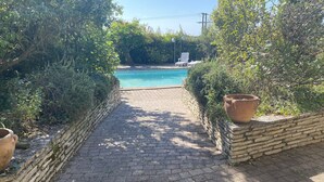 Un chemin pavé mène à une piscine entourée d’une végétation luxuriante et de murs en pierre.