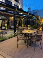 Un área de patio al aire libre con mobiliario de mimbre y una mesa de madera, rodeada por un muro de piedra y una barandilla de vidrio.