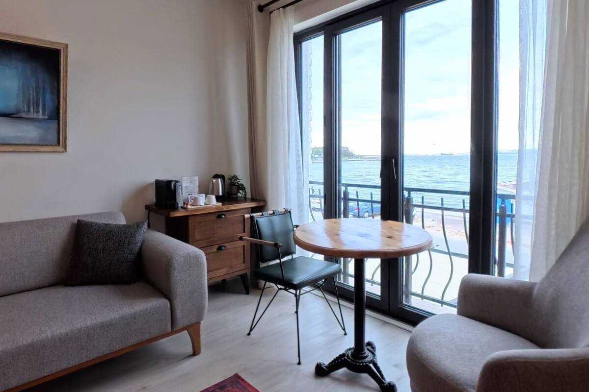 A living room with a sofa, armchair, coffee table, and a red rug. There is a desk with a chair, a window with a view of the sea, and a painting on the wall.
