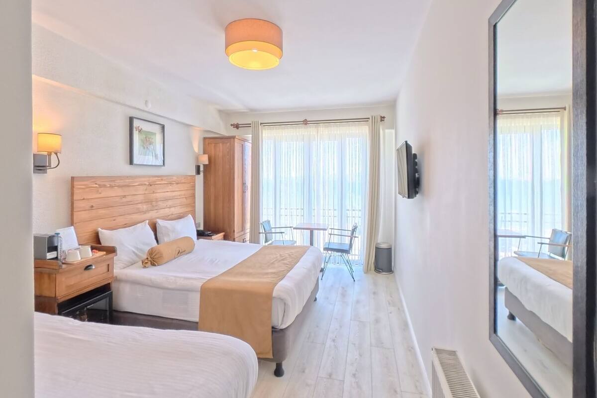 A hotel room with two beds, a wooden headboard, a bedside table with a lamp, a framed picture on the wall, and a large mirror.