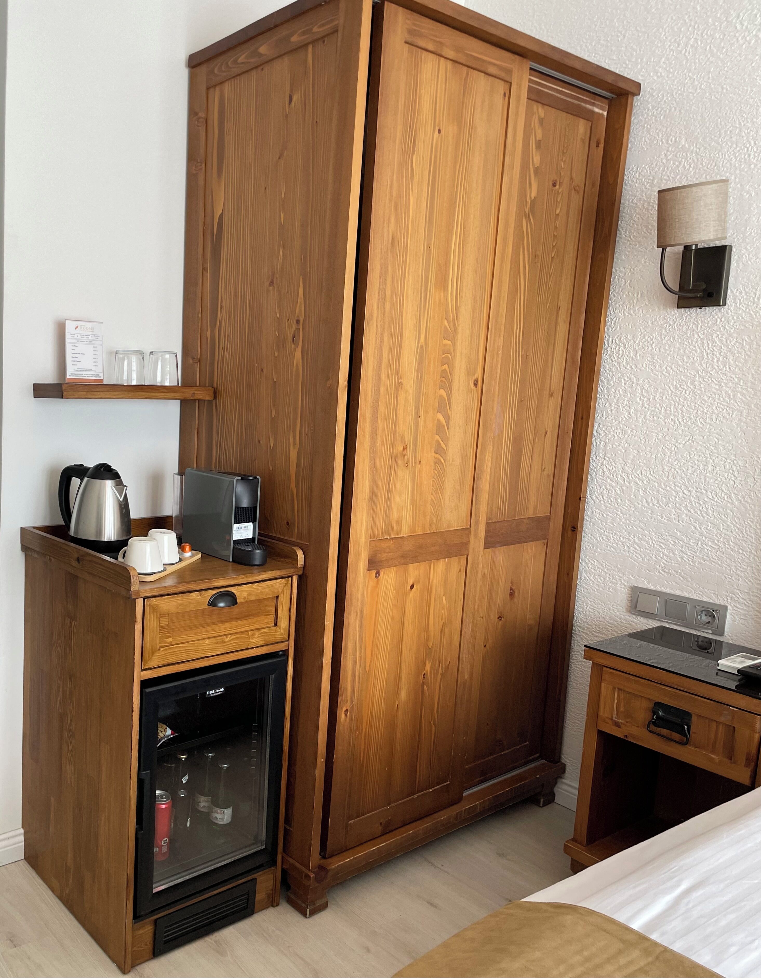 Un armario de madera con minibár de cristal, una mesita de madera con lámpara, y una cama con edredón blanco.