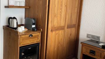 Un armario de madera con minibár de cristal, una mesita de madera con lámpara, y una cama con edredón blanco.
