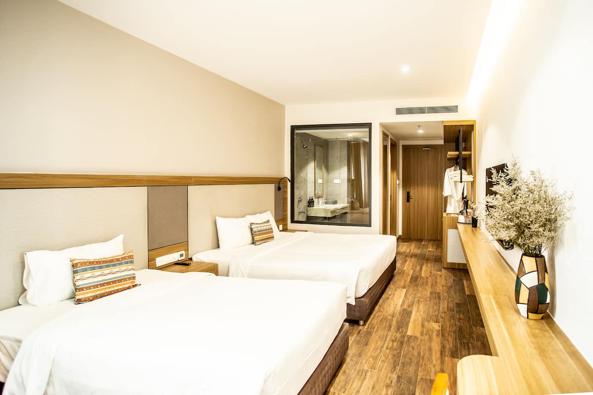A hotel room with two beds, a wooden floor, a mirror, a wooden bench, and a vase with flowers.