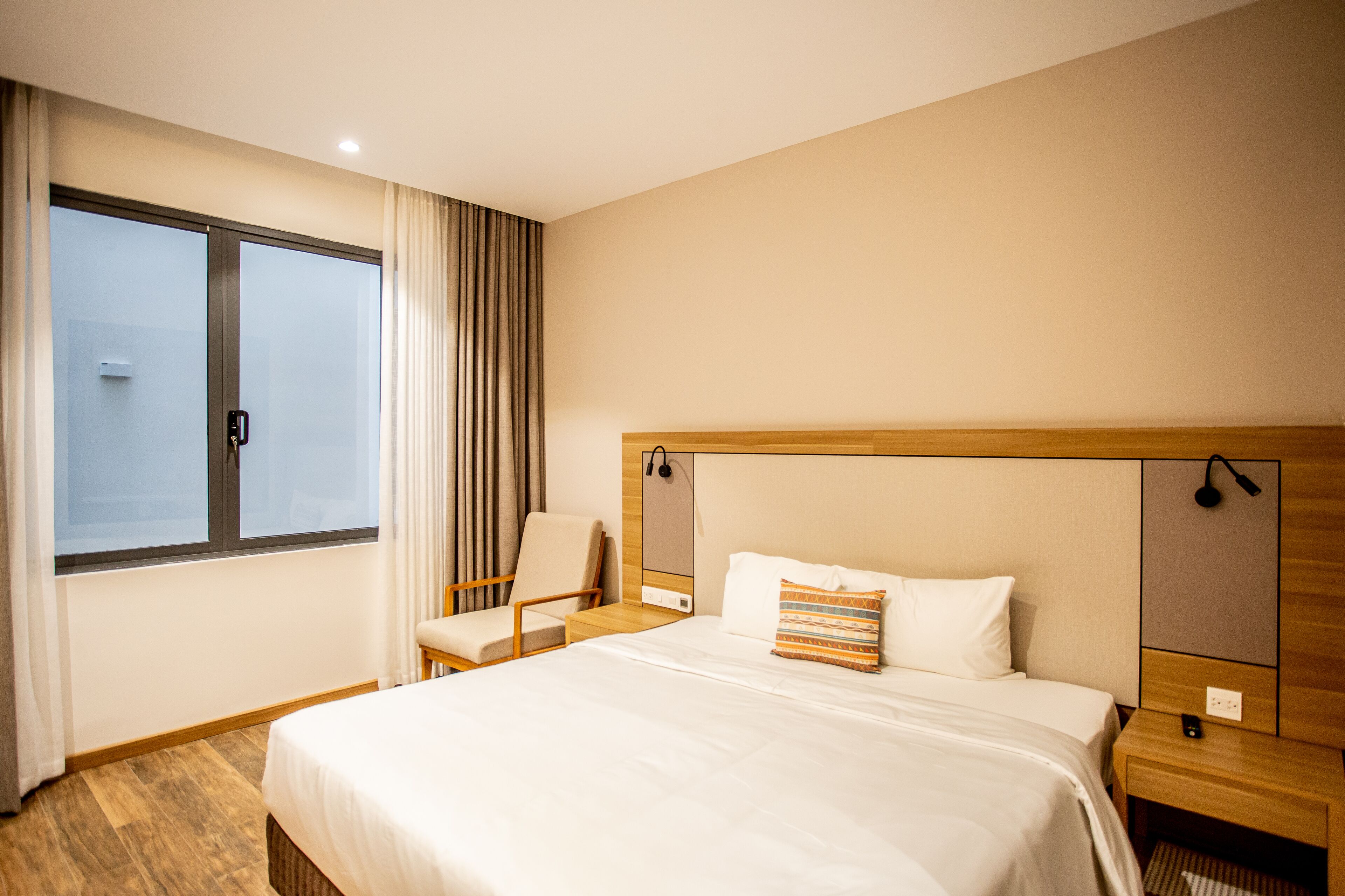 Una habitación de hotel con una cama grande, una silla, una mesita de noche y un ventanal con cortinas.