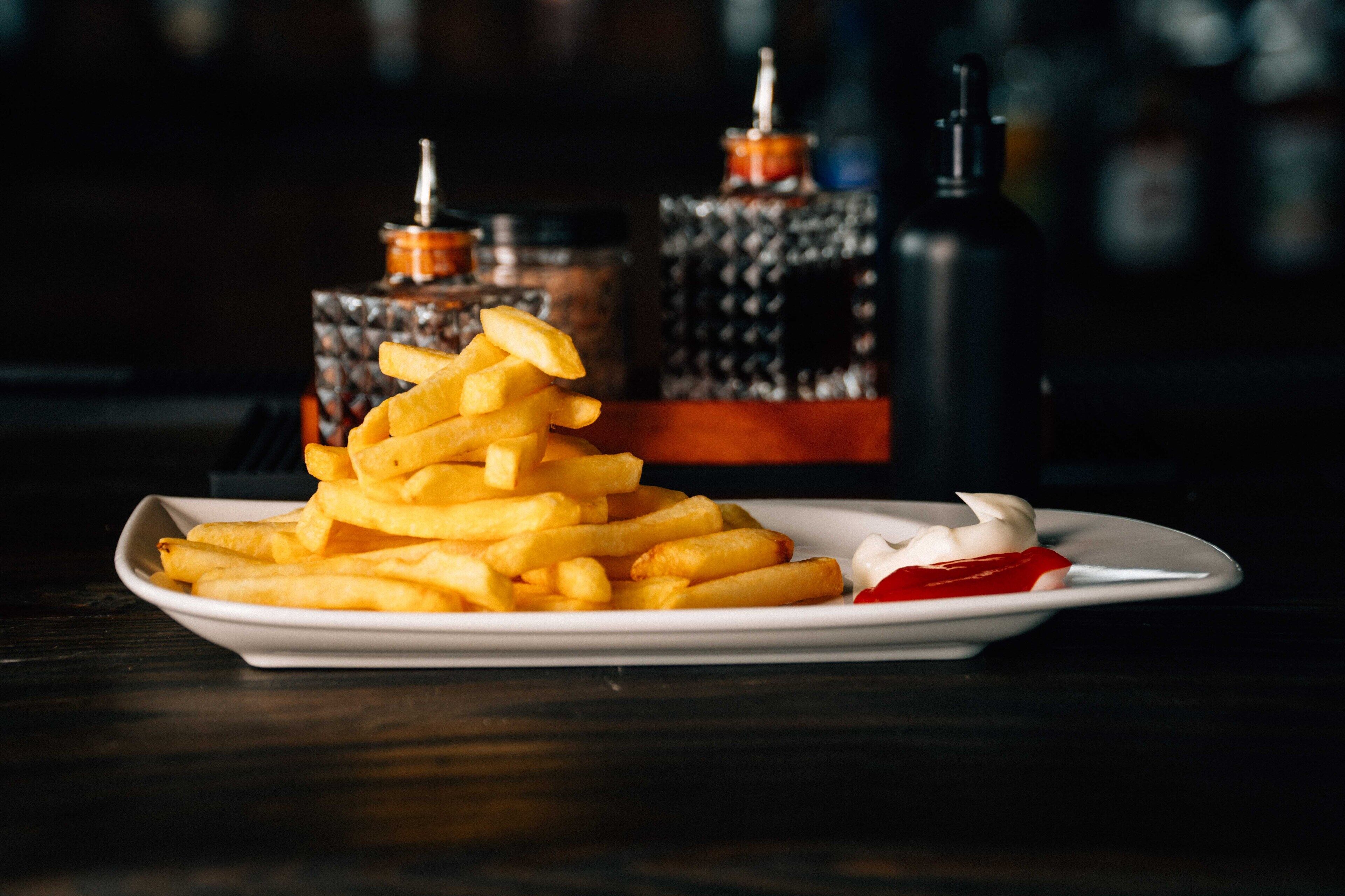 Een bord met goudbruine frieten, ketchup en een lepel mayonaise, voor een donkere achtergrond met onscherpe flesjes saus.