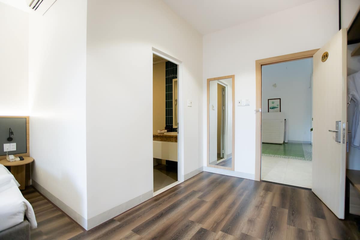 A hotel room with a wooden floor, a bed, a small table with a lamp, and a bathroom visible through an open door.