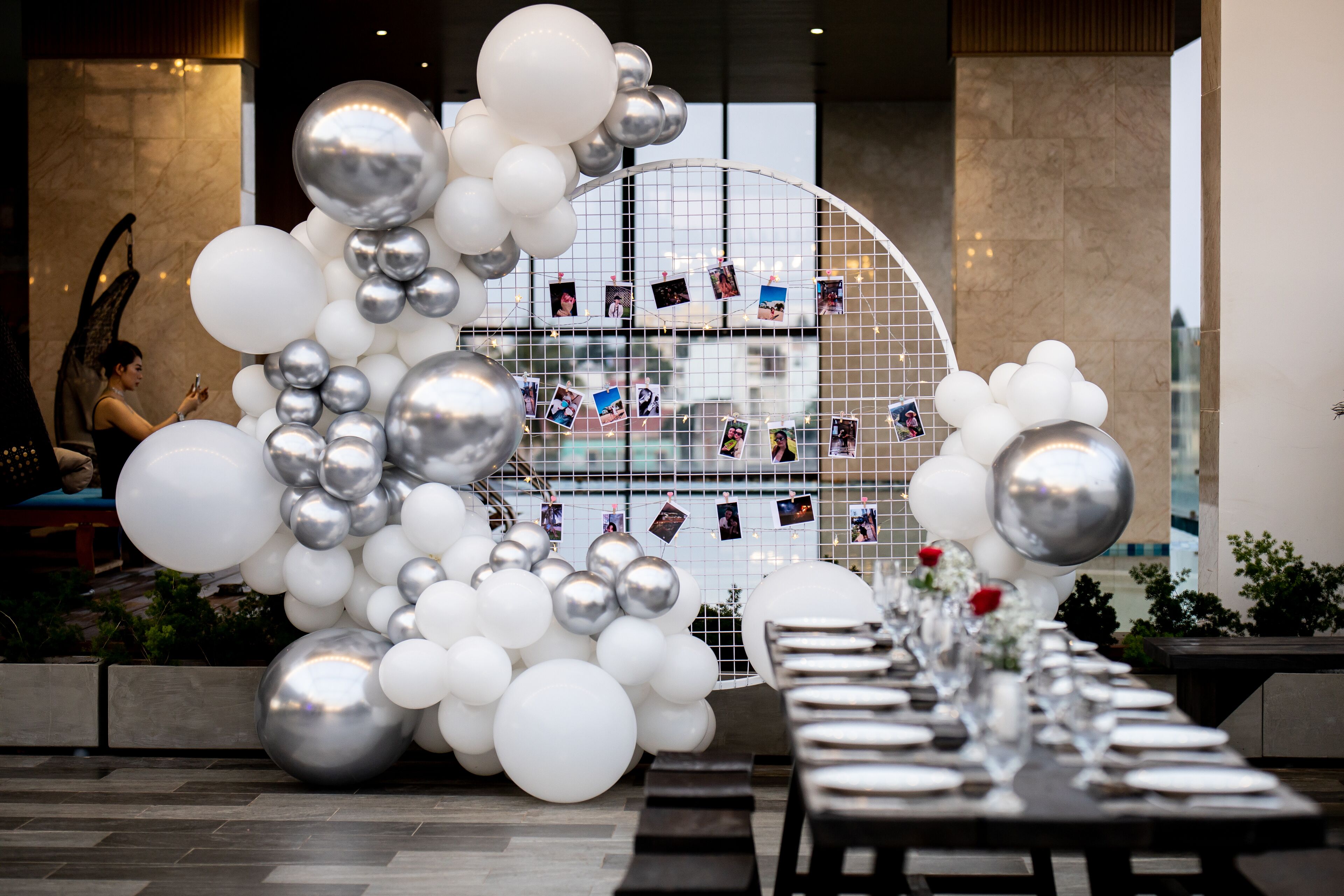 Un espace repas avec une longue table dressée pour un repas, entouré d’une composition décorative de ballons blancs et argentés, et un fond orné d’une grille sur laquelle sont disposées des photos.