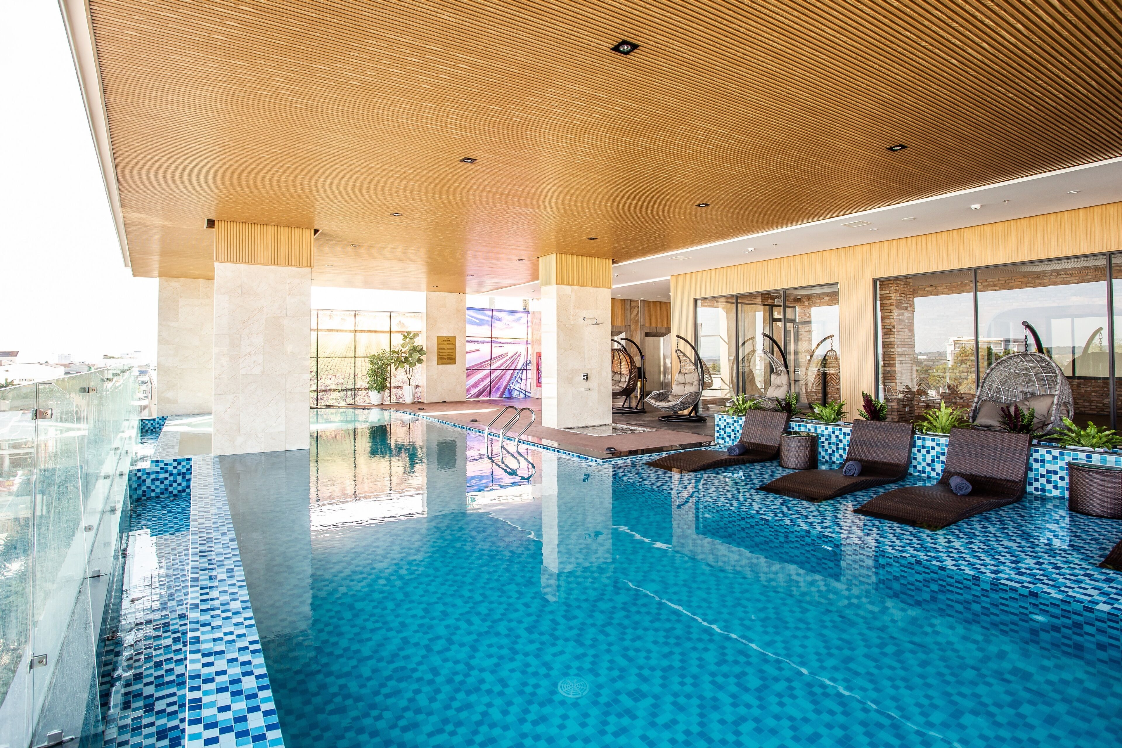 A modern indoor pool with clear glass walls, lounge chairs, and a tiled floor.