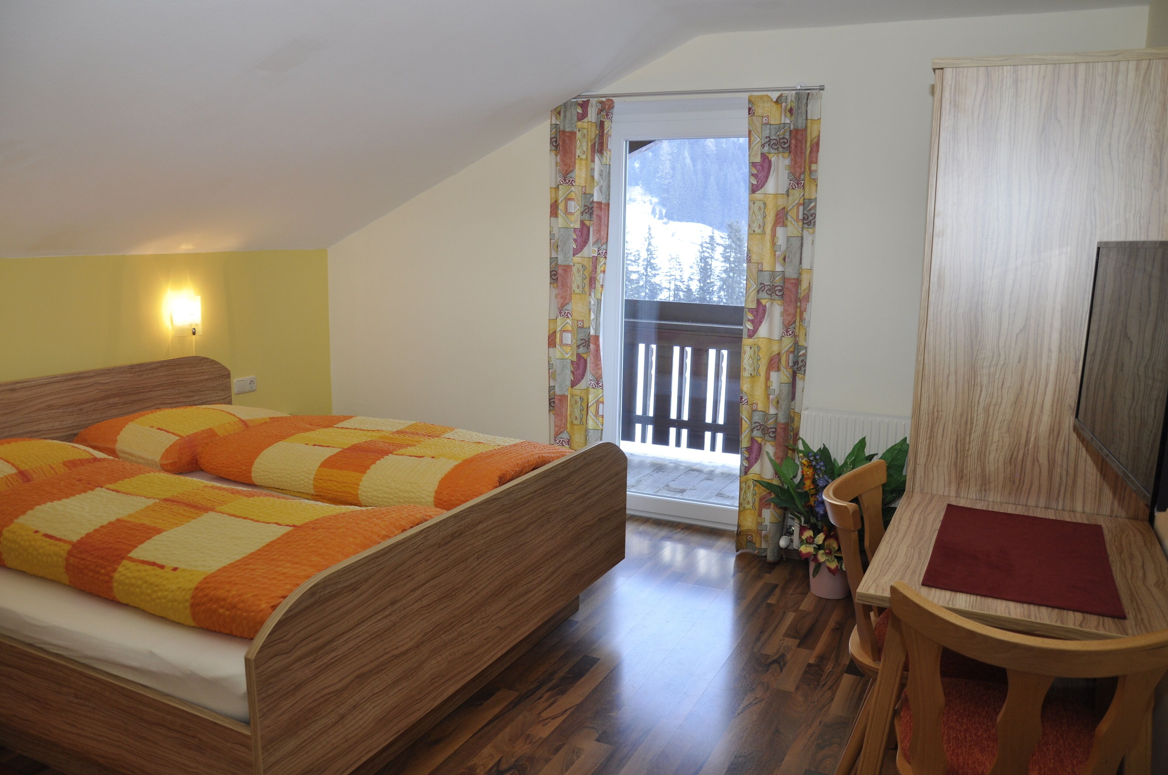 Ein Schlafzimmer mit einem hölzernen Bett, einem kleinen Tisch mit einer roten Unterlage und einem Fenster mit Blick auf eine verschneite Landschaft.