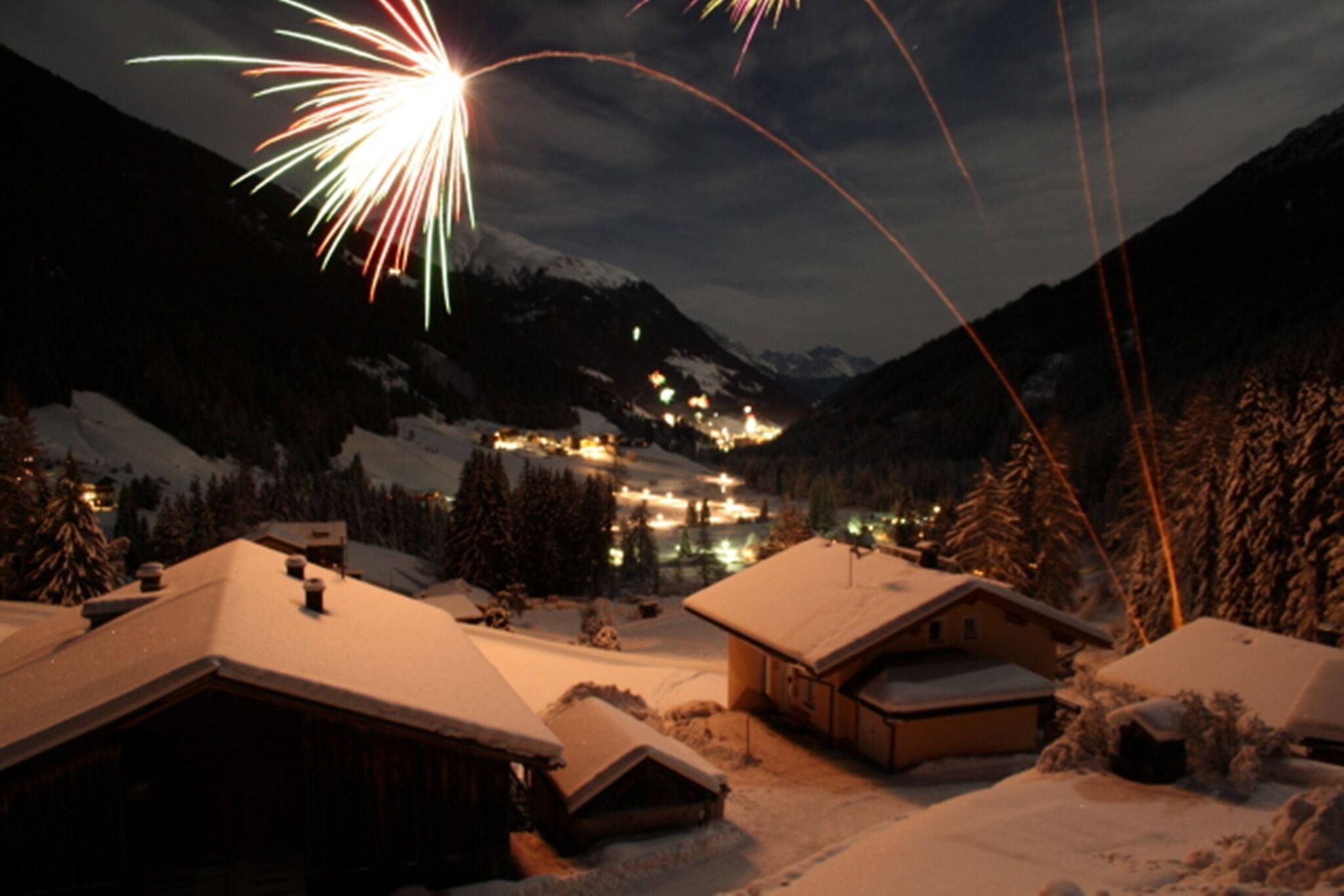 A snowy mountain village with snow-covered houses and a lit-up town in the background.