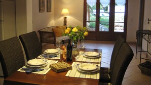 Ein Essbereich mit einem Holztisch, gedeckt für vier Personen, inklusive Tellern, Weingläsern und einer Weinflasche. Korbstühle stehen um den Tisch, und eine Lampe ist an der Wand angebracht. Durch das Fenster ist Grünzeug sichtbar.