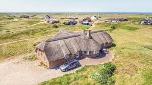 A thatched-roof house with a gravel driveway and a parked car, surrounded by open fields and other similar houses. - Nice home in Harboøre with kitchen (Harboøre)