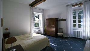 A bedroom with a bed, a wooden wardrobe, a chair, a desk with a lamp, and a window with curtains.