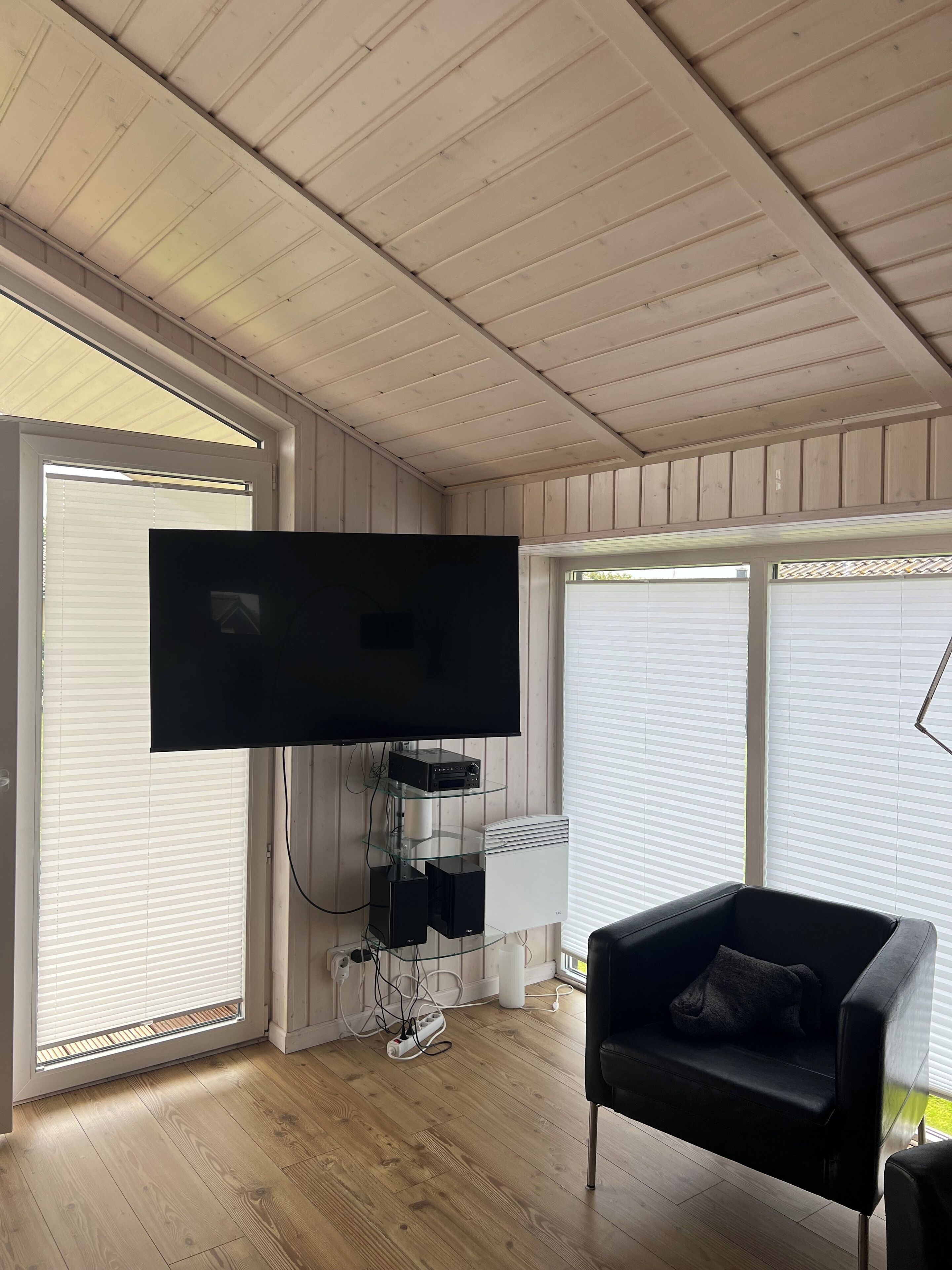 Un salon doté d’un téléviseur à écran plat fixé au mur, d’un canapé en cuir noir et d’une étagère en verre sur laquelle sont posés des appareils électroniques.