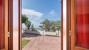 Een open deur naar een balkon met betegelde vloer, een balustrade en uitzicht op bomen en een heldere lucht.