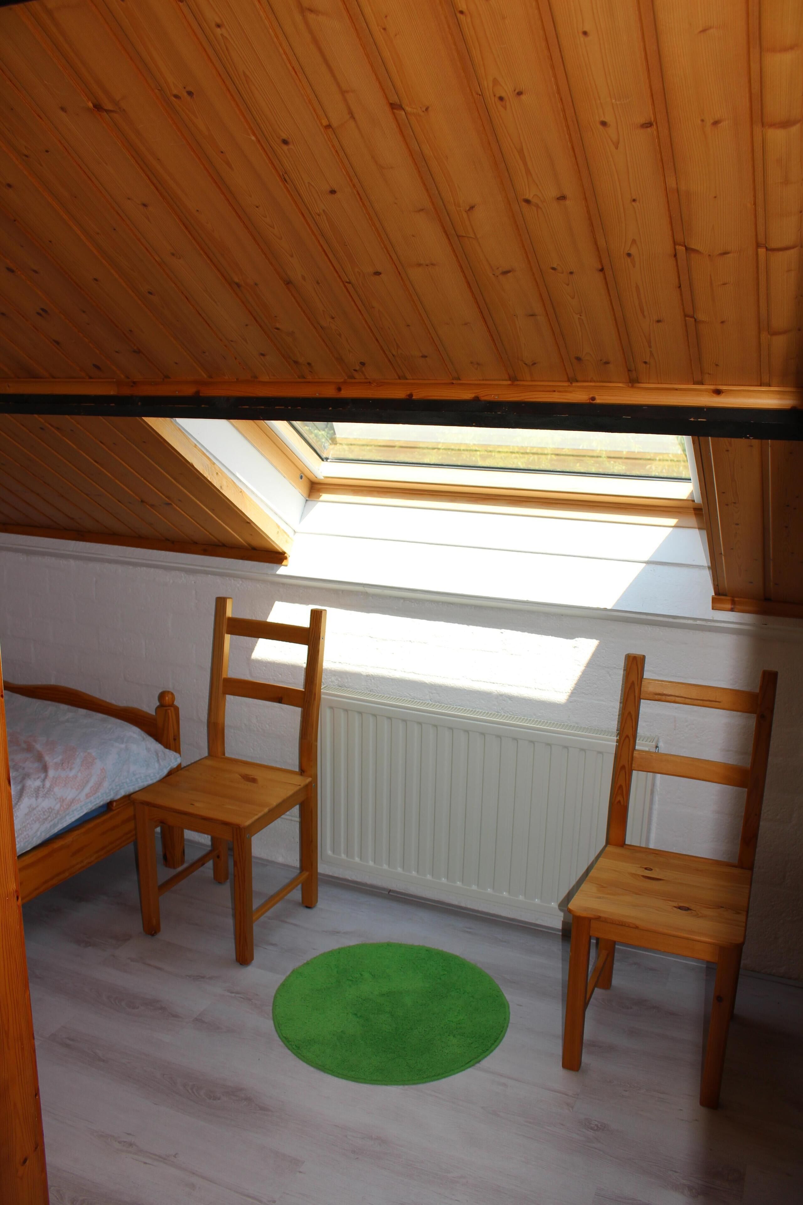 Un pequeño cuarto ático con techo de madera inclinado, un tragaluz, una cama, dos sillas de madera y una alfombra verde.