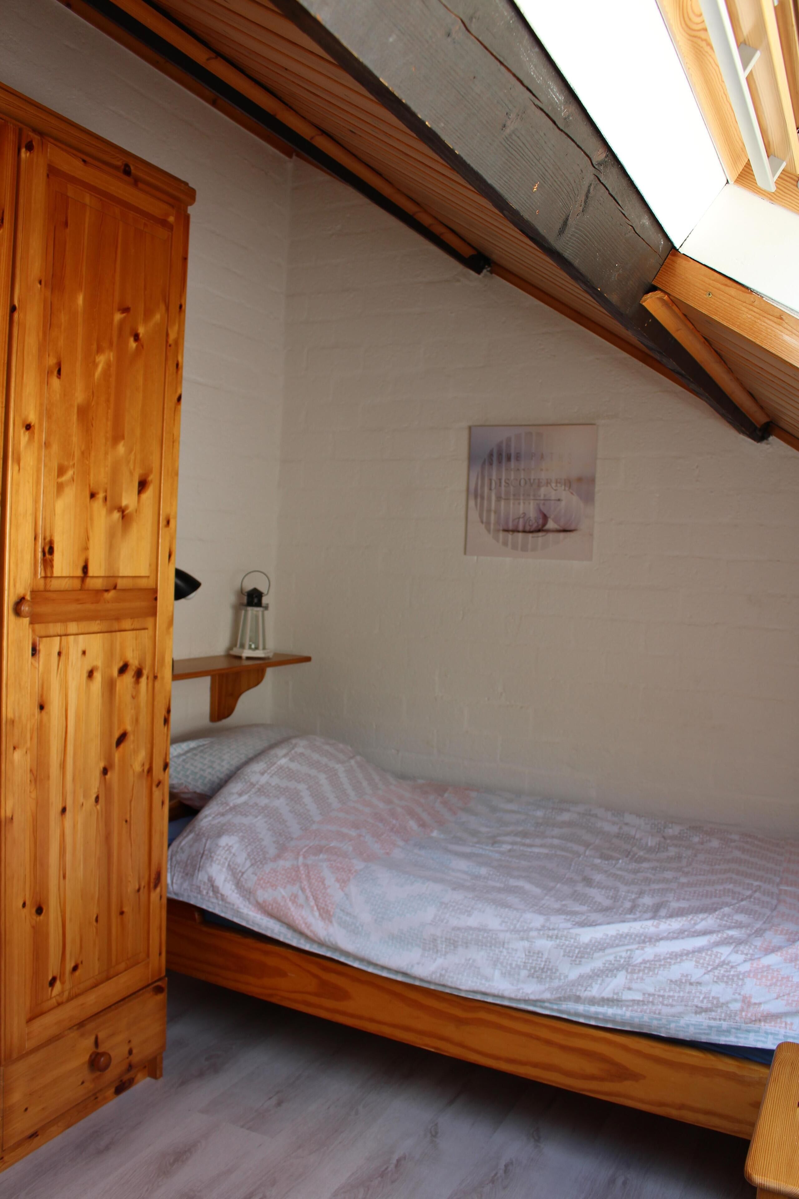 Une chambre avec un lit en bois, une armoire en bois, une petite étagère sur laquelle est posée une lanterne, et un tableau encadré accroché au mur.