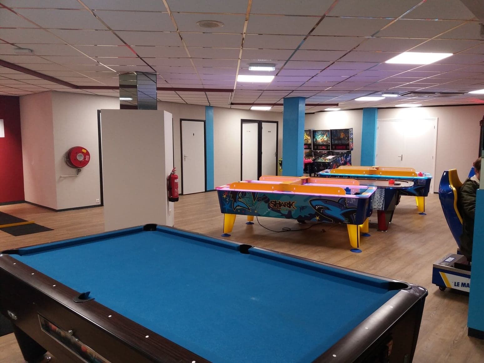 A pool table with a blue felt surface, a fire extinguisher mounted on the wall, and a colorful table tennis table in the background.
