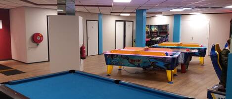 A pool table with a blue felt surface, a fire extinguisher mounted on the wall, and a colorful table tennis table in the background.