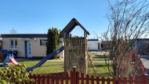 A playground with a slide and climbing structure, a red picket fence, and a building in the background. - Bungalow 58 sqm, 300 m from the beach and subtropical bathing paradise, WLAN (Niedersachsen)