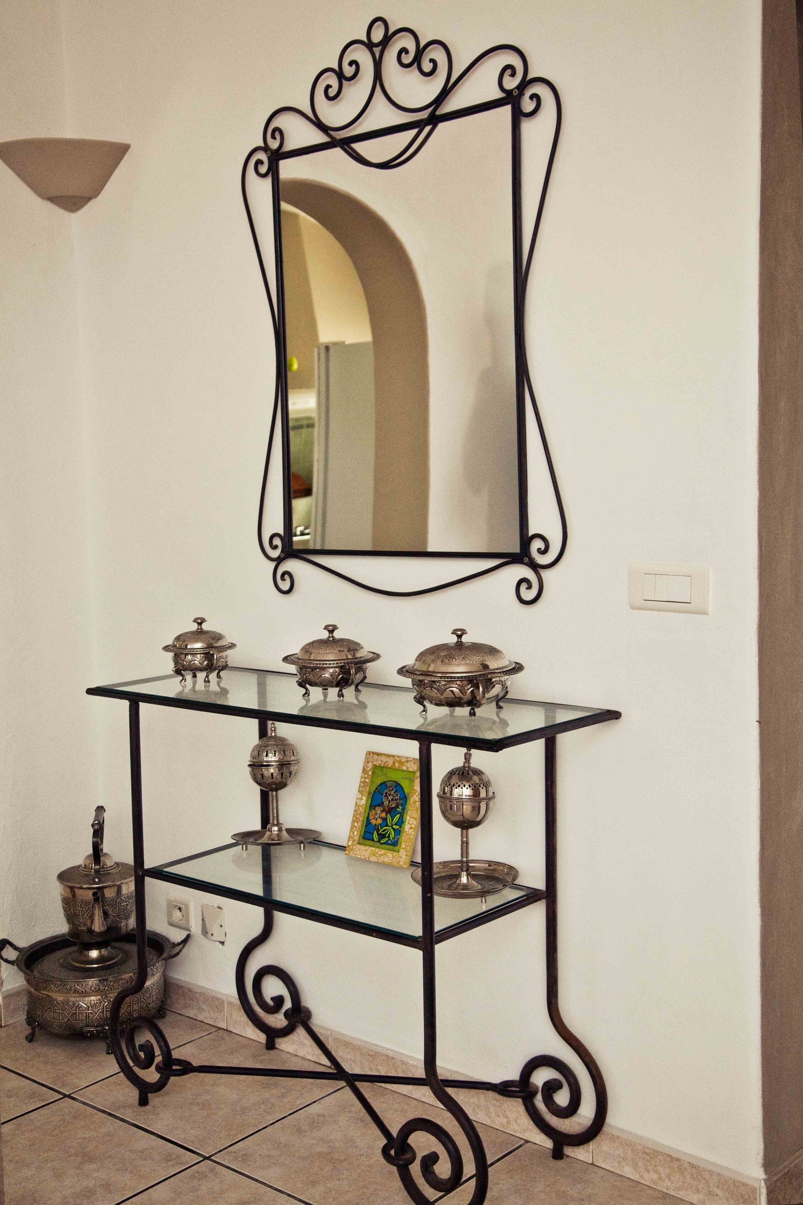 Une console à plateau en verre, dotée de pieds décoratifs en métal et agrémentée d’objets en argent, dont une théière et un tableau encadré, le tout placé devant un mur blanc.