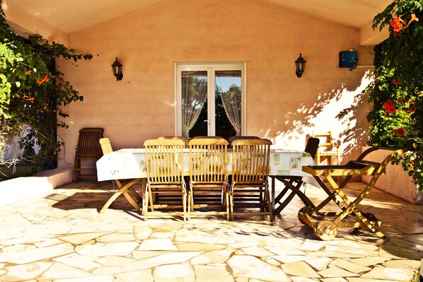 A covered patio with a stone floor, a table with chairs, a small cart, and potted plants. - Magnificent villa with sea view (Zonza)