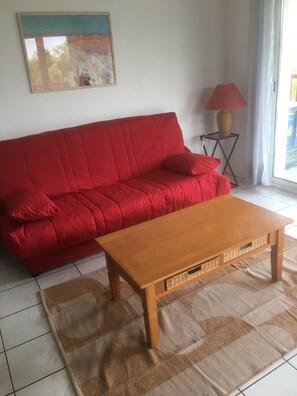 A red sofa with a cushion, a wooden coffee table, a patterned rug, a lamp on a small table, and a painting on the wall.