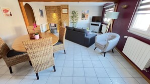 A cozy living area with a round wooden table, wicker chairs, a grey sofa, and a white armchair. There is a potted plant, a radiator, and a TV stand with a flat-screen TV.