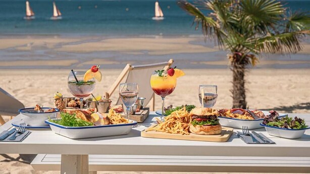 Mesa posta com diversos pratos e bebidas numa praia, com uma palmeira e veleiros ao fundo.