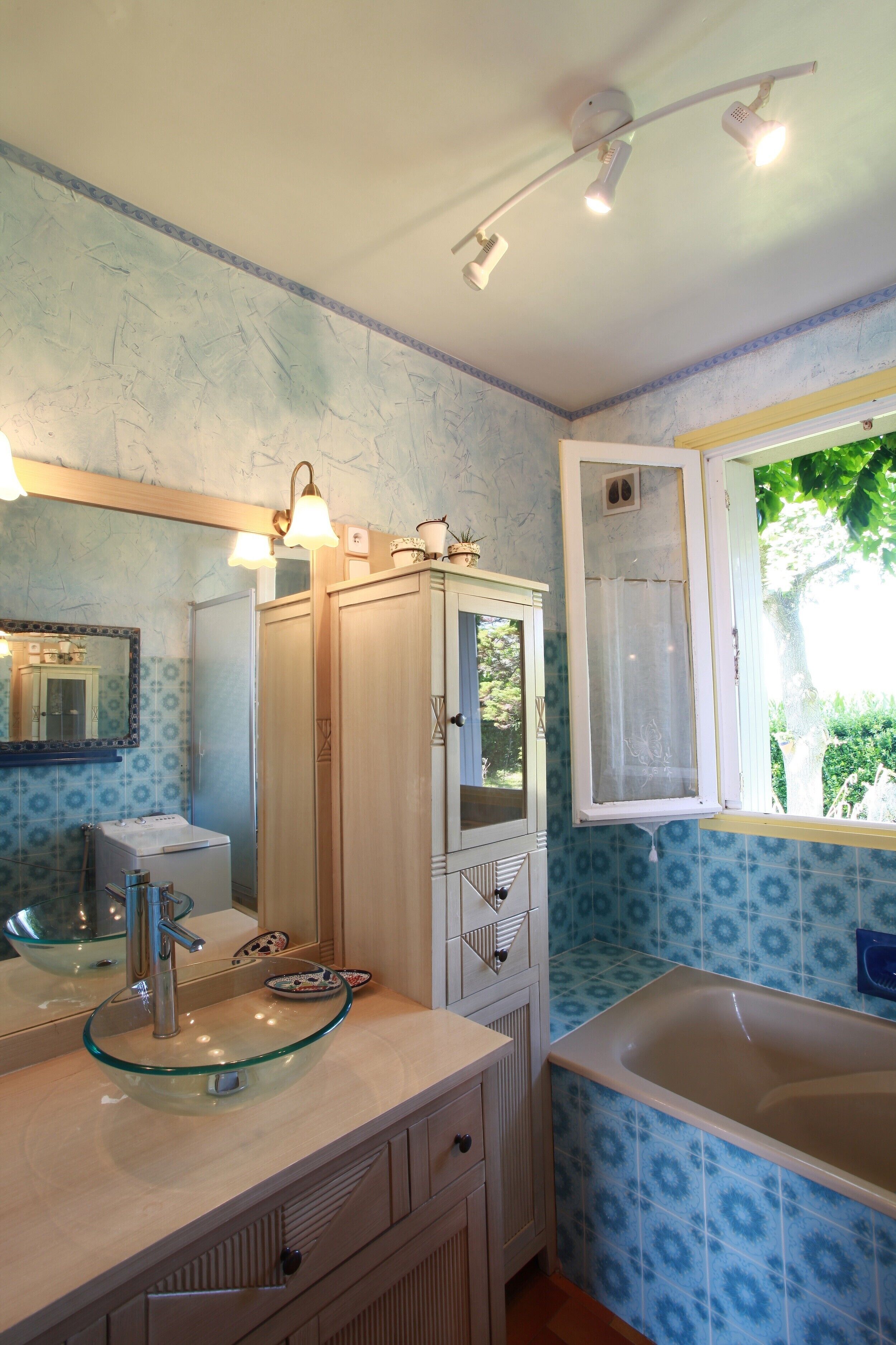 Ein Badezimmer mit einem Glastischwaschbecken, einem Spiegel, einem Schrank, einer Badewanne und einem Fenster mit Blick auf Grünflächen.