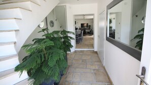 Una escalera con una planta en maceta, un pasillo con piso de baldosas y una puerta que da a una habitación con televisor.