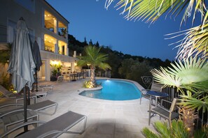 A swimming pool with lounge chairs and umbrellas, surrounded by palm trees and a building with multiple balconies.