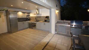 Cocina exterior con electrodomésticos de acero inoxidable, una parrilla y una zona de comedor con mesa y sillas.
