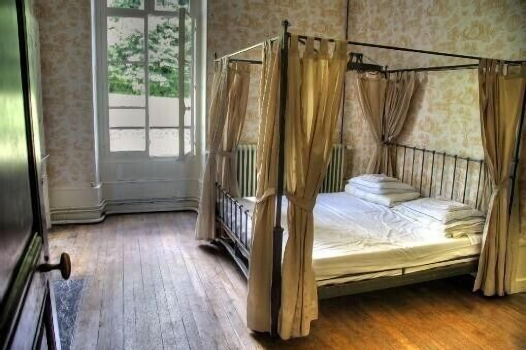 A four-poster bed with curtains, a wooden floor, and a window with a view of greenery.