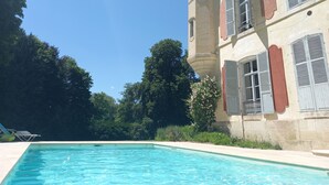 A swimming pool with a clear blue water surface, surrounded by a building with a tower and multiple windows with shutters.