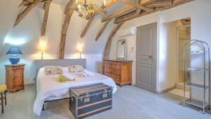 A bedroom with a large bed, wooden beams, a chandelier, a dresser, a mirror, and a metal shelf. - Moulin de Villeverneix and its tower, Neuvic on the banks of the Vern for 6, 8 people (Neuvic)