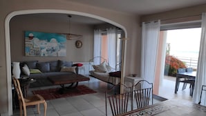 A living room with a sofa, coffee table, and a dining area with a table and chairs. There is a painting on the wall and a view of the outside.
