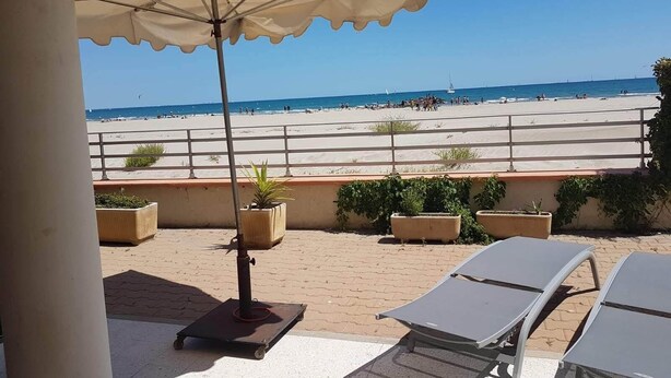 A beach view with a patio area featuring a sun umbrella, lounge chairs, and potted plants.