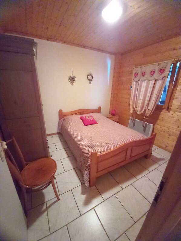 A bedroom with a wooden bed, a chair, a nightstand, and a window with curtains. - CHALET GERARDMER renting the ground floor of 90 m2 plot of 3000m2 (Gérardmer)