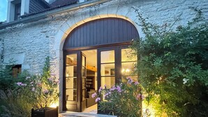 Un edificio de piedra con una amplia entrada en arco, un interior bien iluminado visible a través de puertas de cristal y plantas en macetas a ambos lados de la entrada.