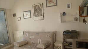 A cozy room with a beige sofa, a small white side table, and a wooden cabinet. There are framed pictures on the wall and a window with a view of the sea.
