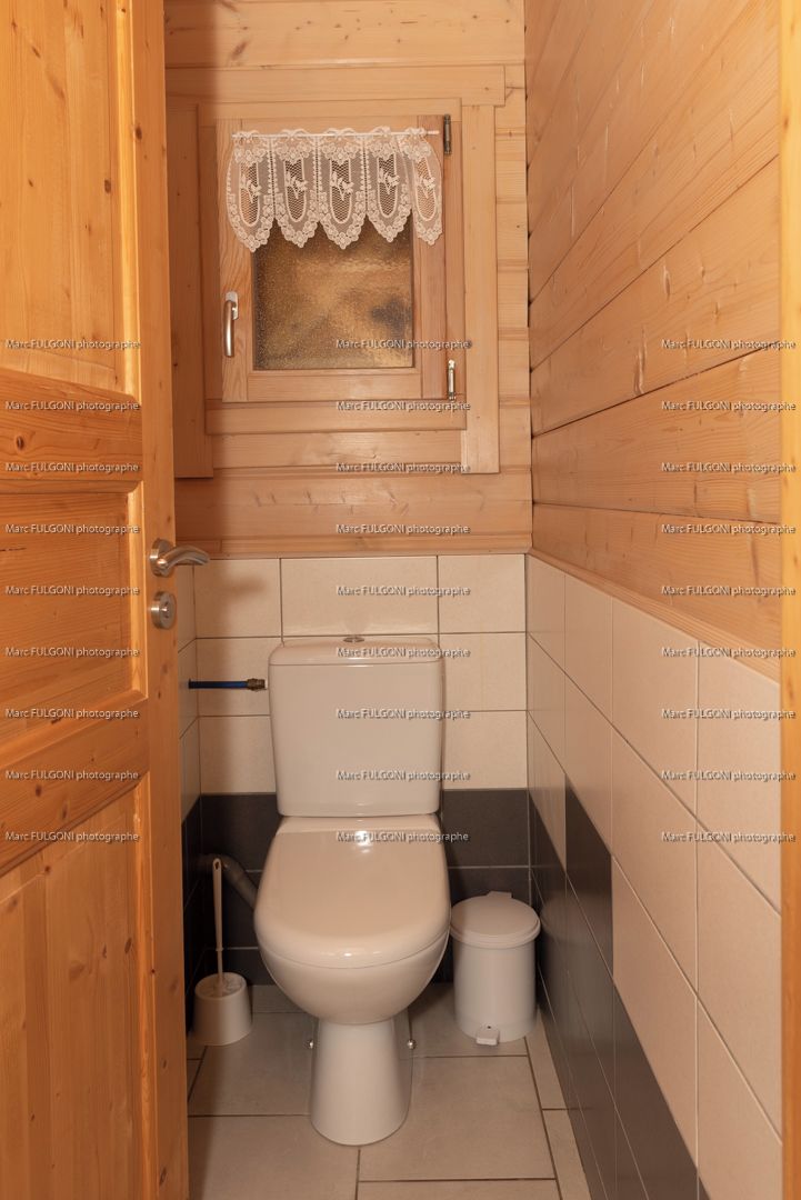 Un baño pequeño con paneles de madera, un inodoro, un ventanal con cortinas de encaje y un cubo de basura.