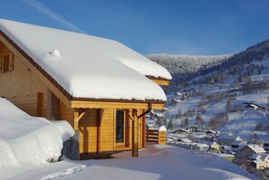 Une cabane en bois dont le toit est recouvert de neige, entourée d’un paysage enneigé et surplombée d’un ciel bleu clair.