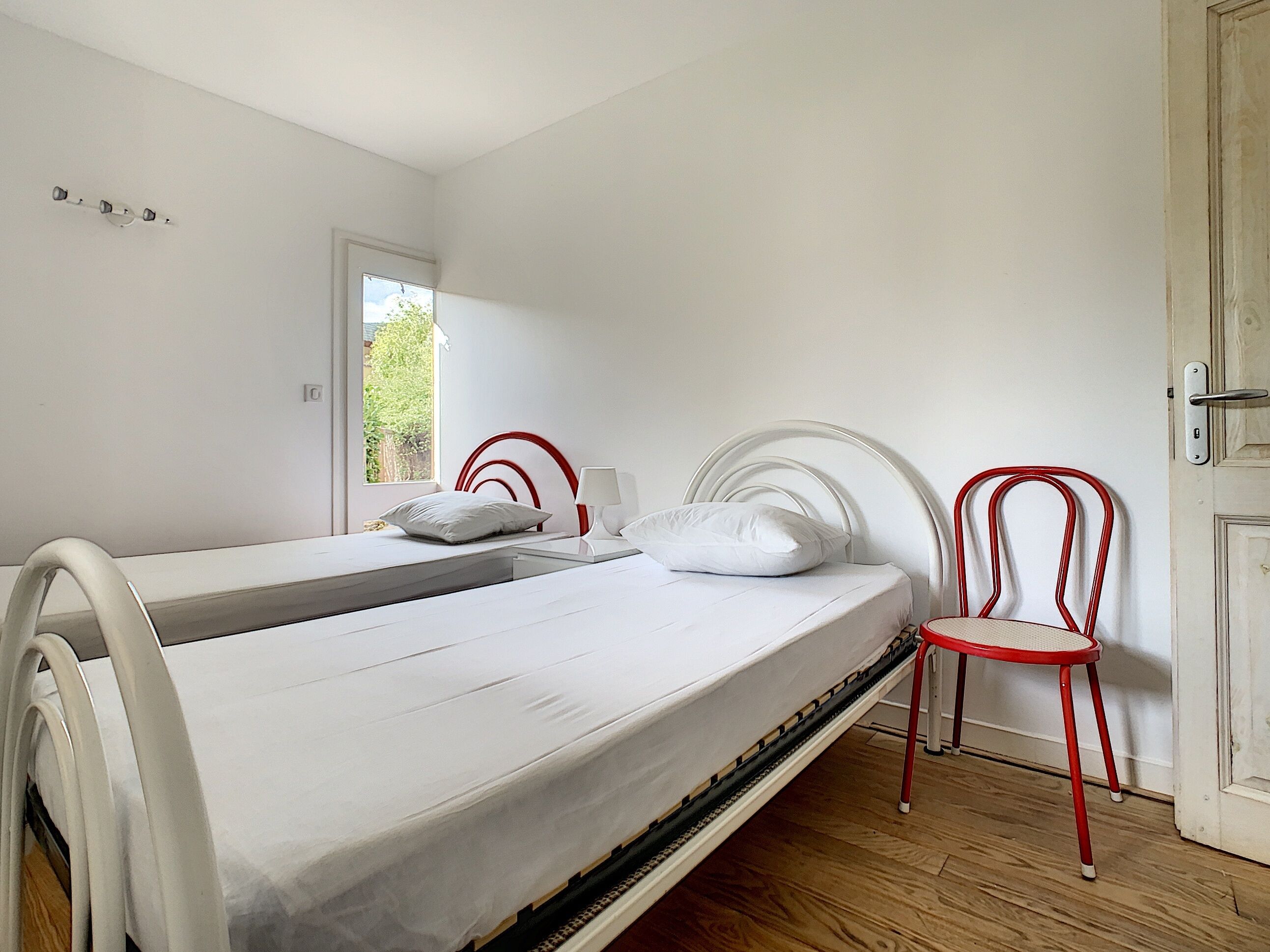 Ein Einzelbett mit weißer Matratze und Kopfteil, ein Nachttisch mit Lampe, ein Fenster mit Blick ins Grüne und ein roter Sessel.