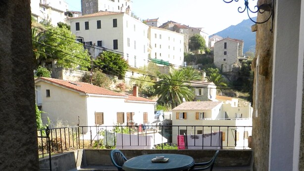 A balcony with a table and chairs, overlooking a hillside with buildings and a mountain in the background. - Apartment of 72 m2 in a house in Corsica (Olmeto)