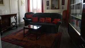 A living room with a dark green sofa, red cushions, a glass coffee table, a wooden side table, and a bookshelf. - Apartment of 72 m2 in a house in Corsica (Olmeto)