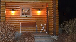 Une cabane en rondins avec deux lampes fixées à l’extérieur sur les murs, un banc et une petite table.