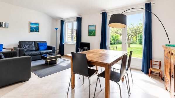 Un salon moderne avec un canapé, une table à manger et des chaises. Il y a des fenêtres avec des rideaux, une lampe et des œuvres d’art accrochées aux murs.