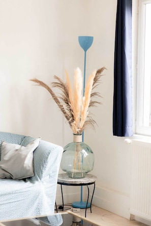 A room with a blue sofa, a glass vase with dried plants, a blue floor lamp, and a window with a dark curtain. - Le Cottage - Ferme de Pontaly (BAILLY)