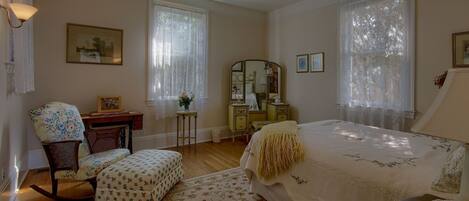 A bedroom with a bed, a chair, a dresser, a mirror, and a window with lace curtains.